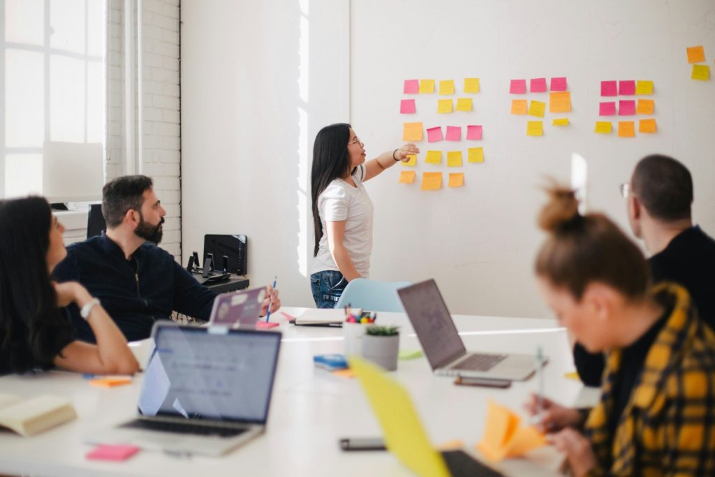 Eine Gruppe von Menschen bei einer Besprechung in einem Büro, während eine Person an einem Whiteboard mit Post-it-Notizen präsentiert. Das Bild symbolisiert die Teamarbeit und die Durchführung von Workshops und Schulungen am Institut für nachhaltige Wassersysteme der Hochschule Hof (inwa).