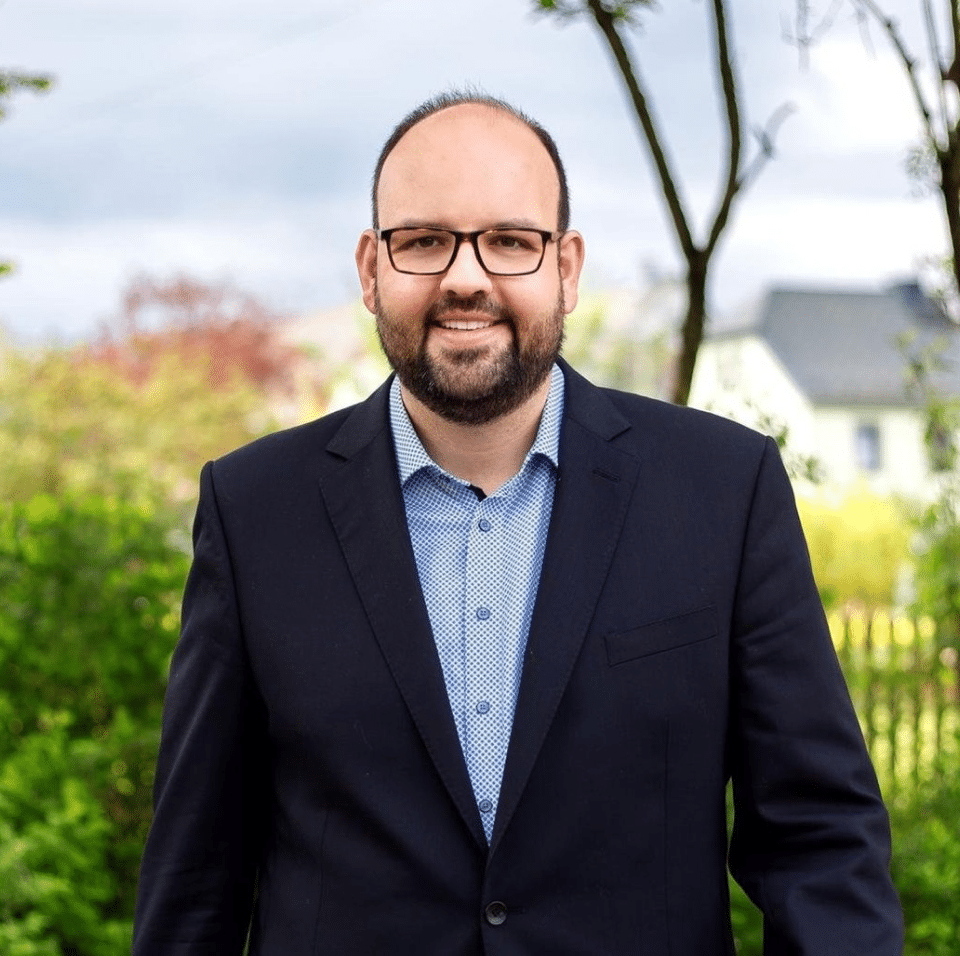 Florian Schaller, erster Bürgermeister der Stadt Schauenstein, lächelt in die Kamera. Im Hintergrund sind Bäume und Häuser zu sehen. Er lobt die Zusammenarbeit mit der Hochschule Hof im Rahmen eines Projekts.